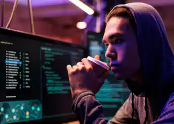 Asian hacker drinking coffee and breaking into database using spyware. Criminal sipping beverage and using malicious software while looking at computer screen in hideout at night