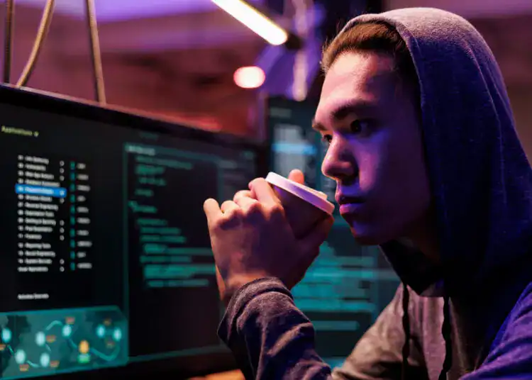 Asian hacker drinking coffee and breaking into database using spyware. Criminal sipping beverage and using malicious software while looking at computer screen in hideout at night