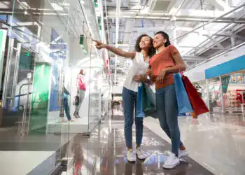 Two smiling black beautiful girls looking at shop window, holding bags and standing in shopping mall. One girl is pointing at something.