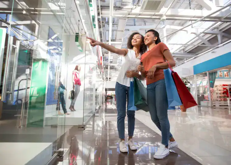 Two smiling black beautiful girls looking at shop window, holding bags and standing in shopping mall. One girl is pointing at something.