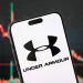 CHONGQING, CHINA - MAY 11: In this photo illustration, the logo of Under Armour, Inc. is displayed on a smartphone screen, with the company's latest stock market chart in the background, reflecting investor sentiment and recent trading activity, on May 11, 2025, in Chongqing, China. Under Armour is an American sportswear and athletic performance apparel company known for its innovative moisture-wicking fabrics and high-performance gear. (Photo illustration by Cheng Xin/Getty Images)