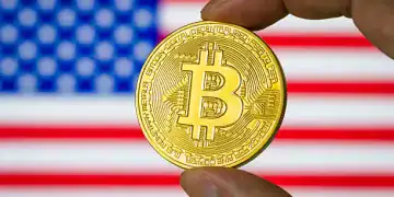 CHONGQING, CHINA - JULY 21: In this photo illustration, a person holds a physical representation of Bitcoin (BTC) in front of a screen displaying the national flag of the United States of America, reflecting the increasing relevance of cryptocurrency in the American financial and regulatory landscape on July 21, 2025 in Chongqing, China. (Photo illustration by Cheng Xin/Getty Images)