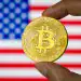 CHONGQING, CHINA - JULY 21: In this photo illustration, a person holds a physical representation of Bitcoin (BTC) in front of a screen displaying the national flag of the United States of America, reflecting the increasing relevance of cryptocurrency in the American financial and regulatory landscape on July 21, 2025 in Chongqing, China. (Photo illustration by Cheng Xin/Getty Images)