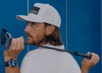 alt A man wearing a white cap and shirt holds a golf club over his shoulder. He is looking back over his left shoulder. He also wears a smartwatch on his left wrist. Blue lockers are visible in the background.