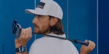 alt A man wearing a white cap and shirt holds a golf club over his shoulder. He is looking back over his left shoulder. He also wears a smartwatch on his left wrist. Blue lockers are visible in the background.