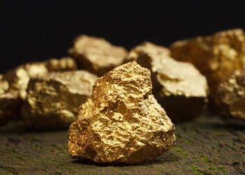 Lingotes y monedas de oro sobre una mesa junto a gráficos de cotización de metales preciosos