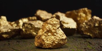 Lingotes y monedas de oro sobre una mesa junto a gráficos de cotización de metales preciosos
