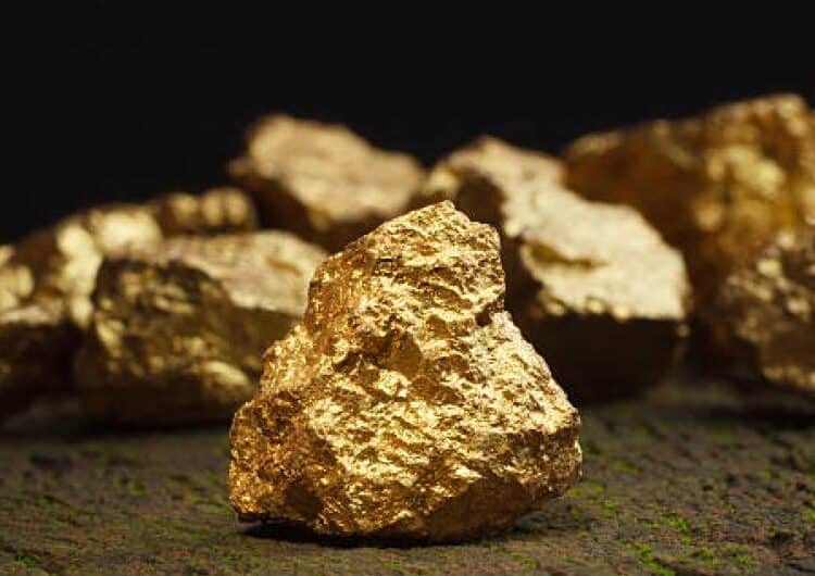 Lingotes y monedas de oro sobre una mesa junto a gráficos de cotización de metales preciosos