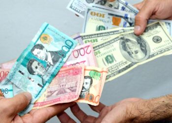 Billetes de dólares y pesos dominicanos sobre una mesa, con gráfica de tipo de cambio al fondo