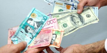 Billetes de dólares y pesos dominicanos sobre una mesa, con gráfica de tipo de cambio al fondo