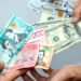 Billetes de dólares y pesos dominicanos sobre una mesa, con gráfica de tipo de cambio al fondo