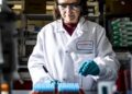 Científico sosteniendo una dosis de vacuna ARNm en laboratorio farmacéutico con equipo de investigación oncológica al fondo