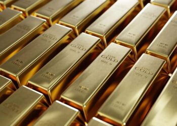 Barras de oro apiladas sobre una superficie reflectante con gráficos de precios al fondo
