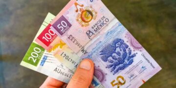 Billetes de pesos mexicanos y dólares estadounidenses sobre una mesa, con gráfica de tipo de cambio al fondo