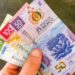 Billetes de pesos mexicanos y dólares estadounidenses sobre una mesa, con gráfica de tipo de cambio al fondo