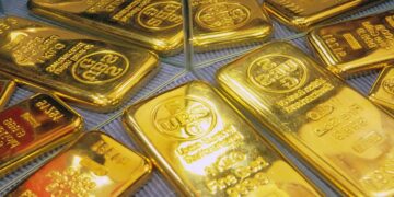 Lingotes de oro apilados sobre una superficie reflejante con gráfico de cotizaciones al fondo