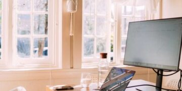 Persona trabajando frente a una computadora de escritorio en una oficina moderna junto a una laptop cerrada sobre el escritorio