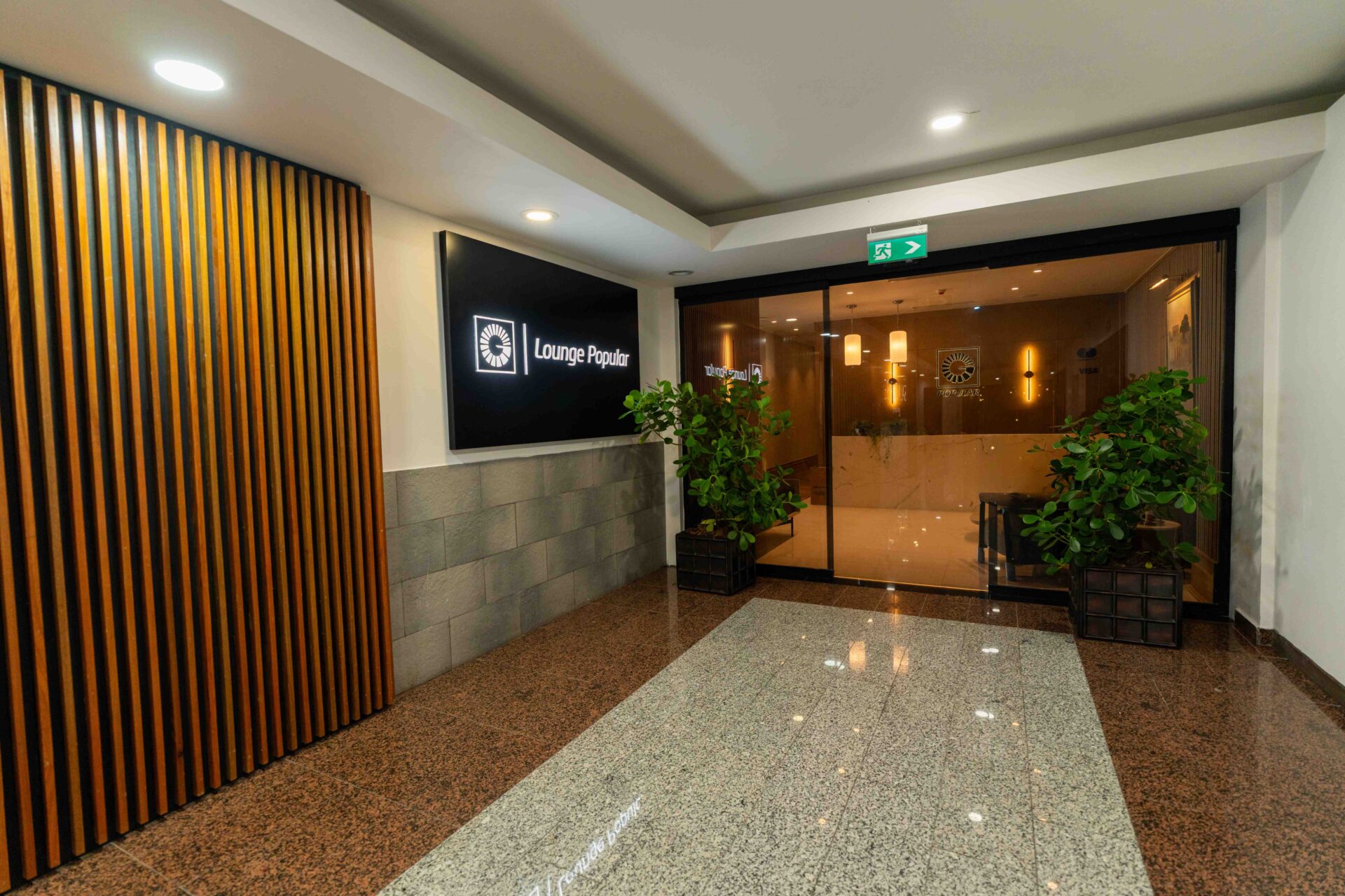 Entrada del Lounge Popular con letrero iluminado, plantas verdes y puertas de vidrio.