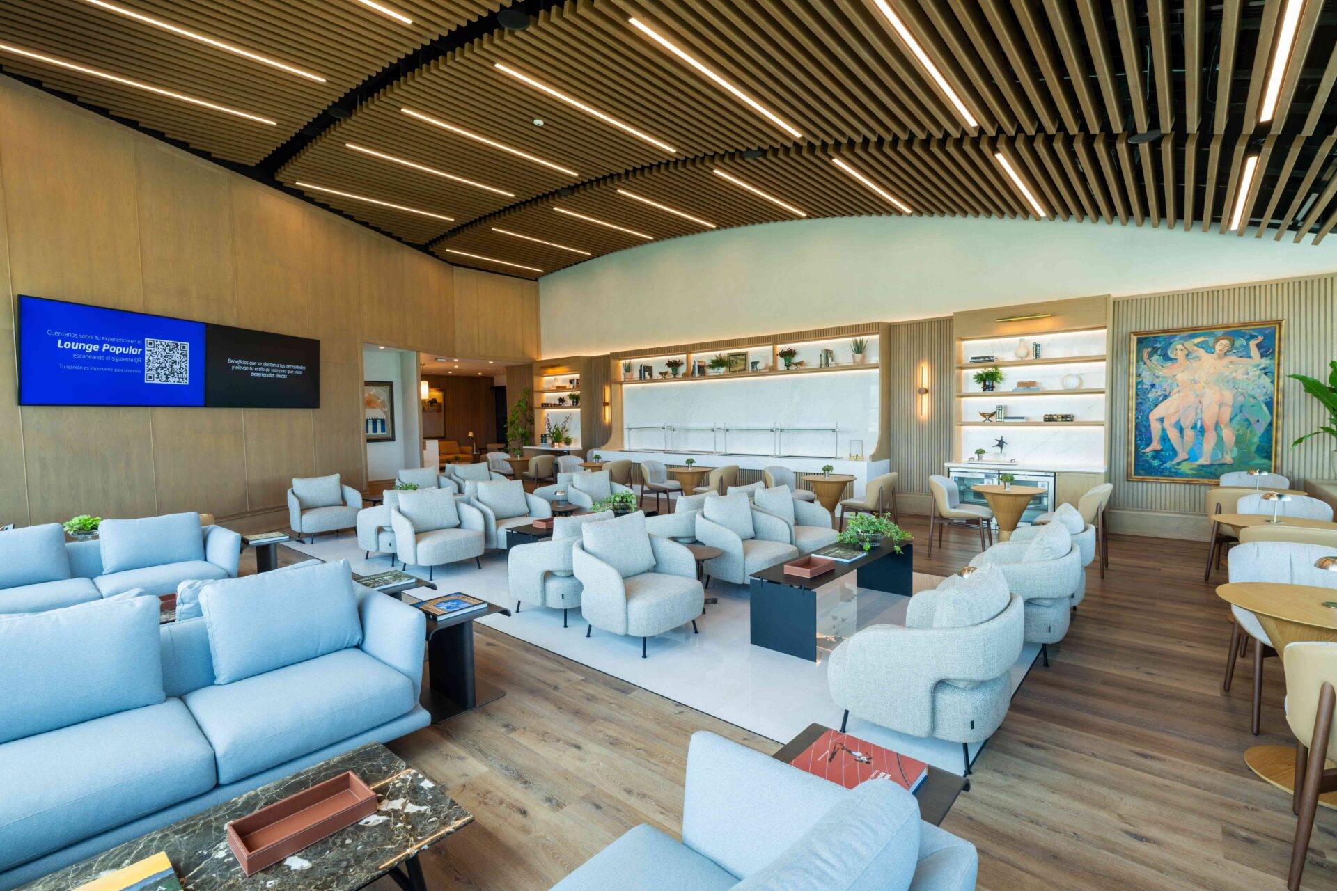 Interior moderno del Lounge Popular con sofás azules, sillones grises, piso de madera y estantería iluminada al fondo.