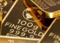 Lingotes de oro apilados sobre superficie metálica con gráfico de precios al fondo