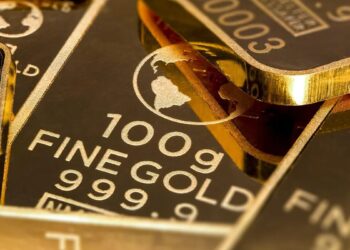 Lingotes de oro apilados sobre superficie metálica con gráfico de precios al fondo