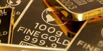 Lingotes de oro apilados sobre superficie metálica con gráfico de precios al fondo