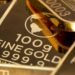 Lingotes de oro apilados sobre superficie metálica con gráfico de precios al fondo