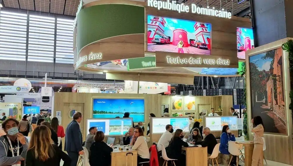 Stand de República Dominicana en una feria internacional de turismo con visitantes reunidos en mesas.