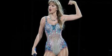 Taylor Swift en el escenario durante la gira The Eras Tour, sosteniendo una guitarra rosa con botas plateadas y luces moradas de fondo