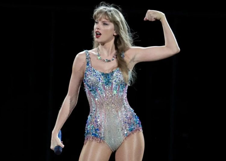 Taylor Swift en el escenario durante la gira The Eras Tour, sosteniendo una guitarra rosa con botas plateadas y luces moradas de fondo