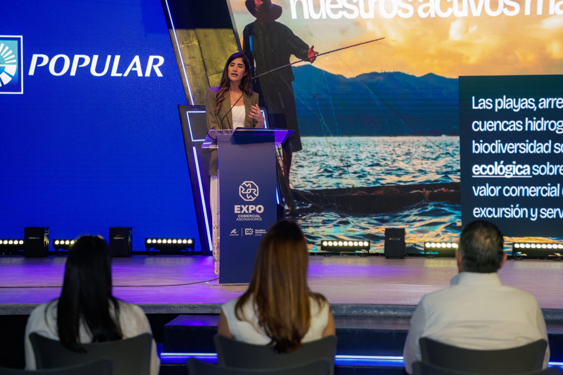 Una ejecutiva presenta en el Expo Comercial Asonahores ante el logo del Banco Popular.