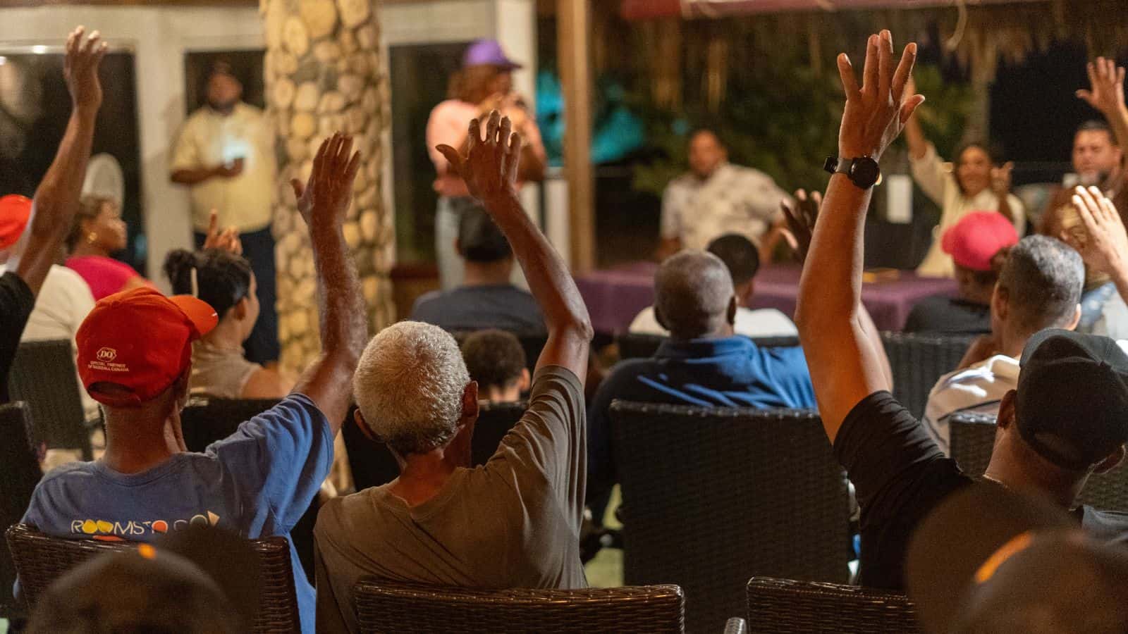 Asistentes levantan la mano durante una asamblea política en un recinto cubierto.
