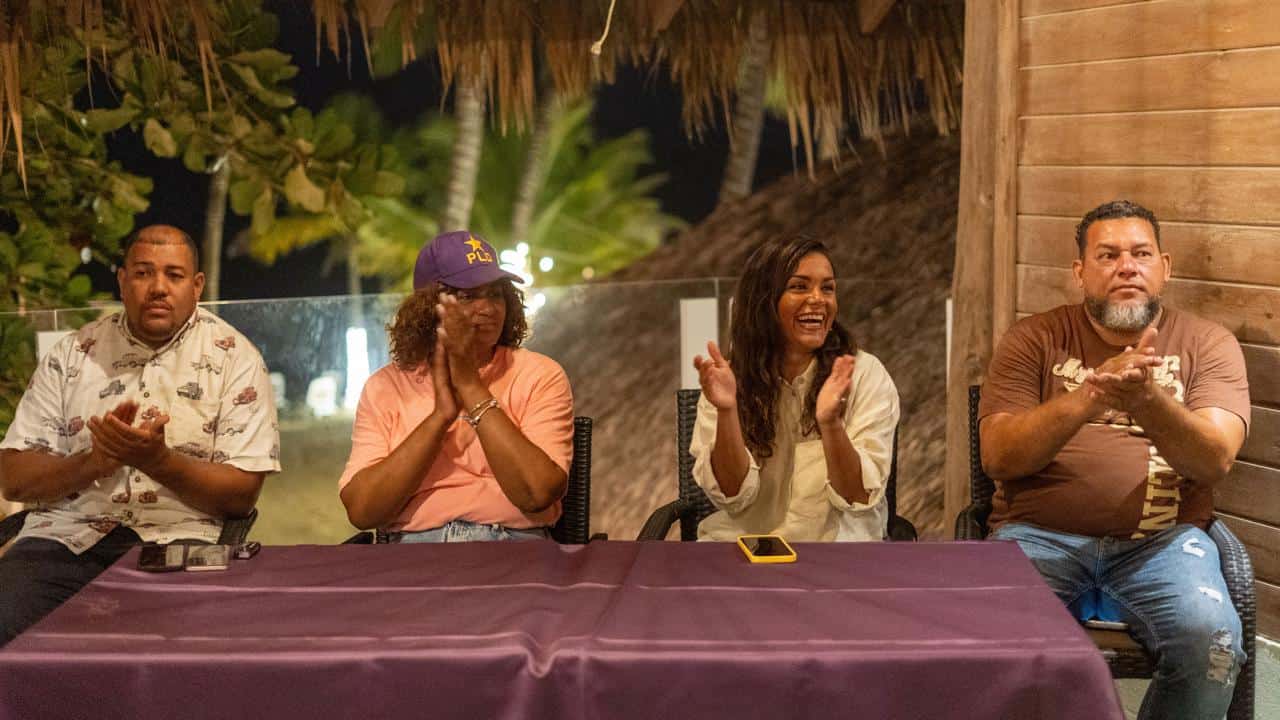 Cuatro personas aplauden sentadas tras una mesa con mantel morado en un evento nocturno al aire libre del PLD.