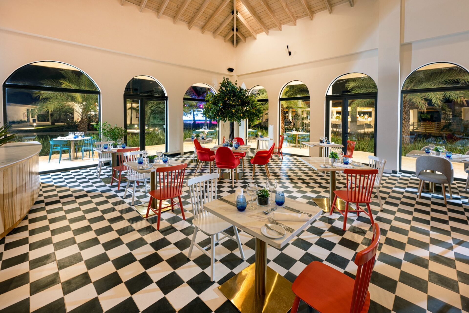 Interior de un restaurante con sillas rojas y blancas sobre piso de ajedrez en blanco y negro, mesas de mármol y arcos con