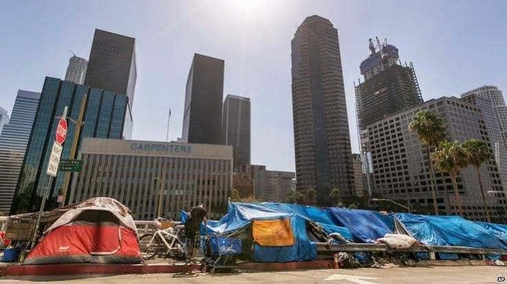 Personas sin hogar en carpas en una calle de Los Ángeles, California, con edificios de apartamentos al fondo