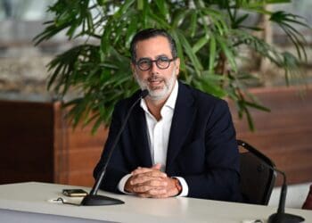 Un hombre con gafas y blazer oscuro habla ante un micrófono en una mesa.