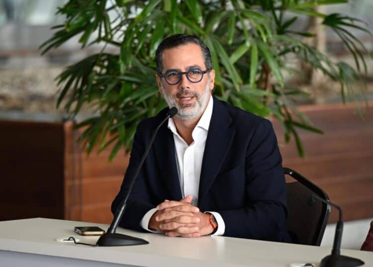 Un hombre con gafas y blazer oscuro habla ante un micrófono en una mesa.