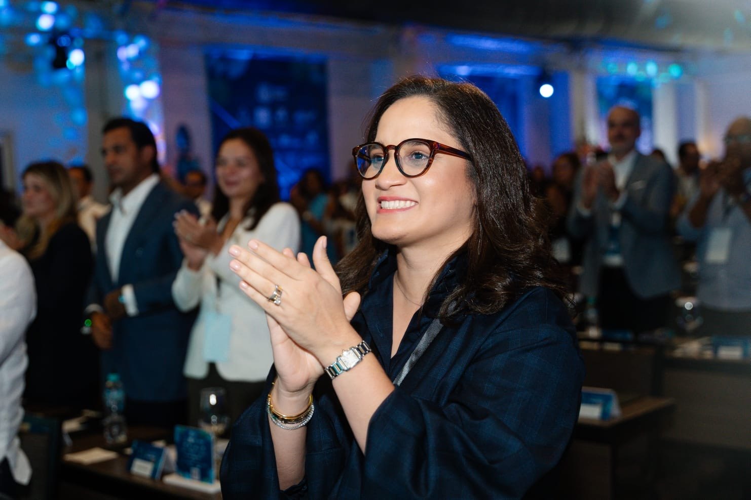 Una ejecutiva con gafas aplaude sonriente en un congreso empresarial.