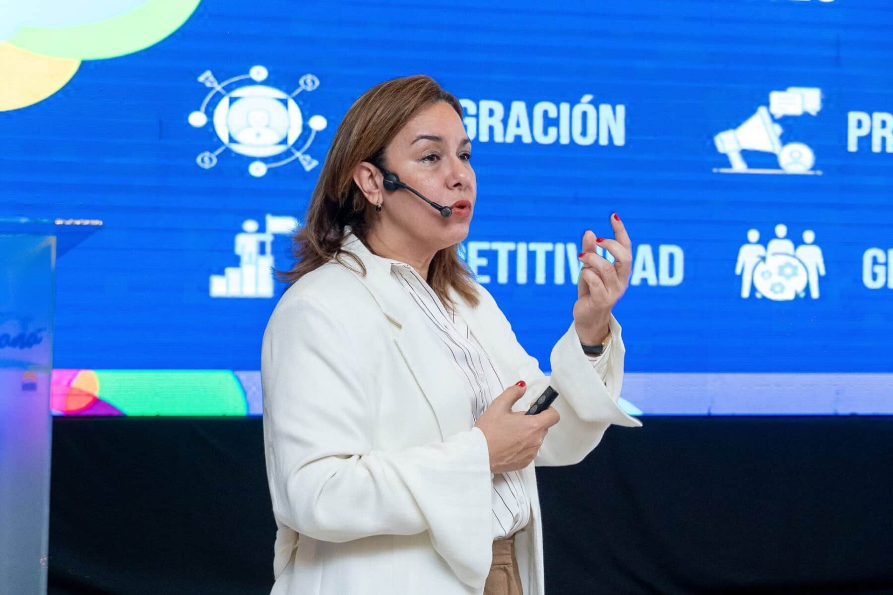 Una ponente con micrófono de diadema expone frente a una pantalla azul con términos de competitividad e integración.